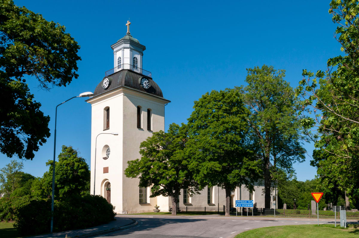 En ljus bild tagen utomhus från en gata som svänger förbi en kyrka i ljus färg med ett koppartak och en guldkors. Omkring kyrkan syns grönskande träd, buskar och en gräsmatta. En gatlykta, en anvisningsskylt och en väjningspliktsskylt syns också.