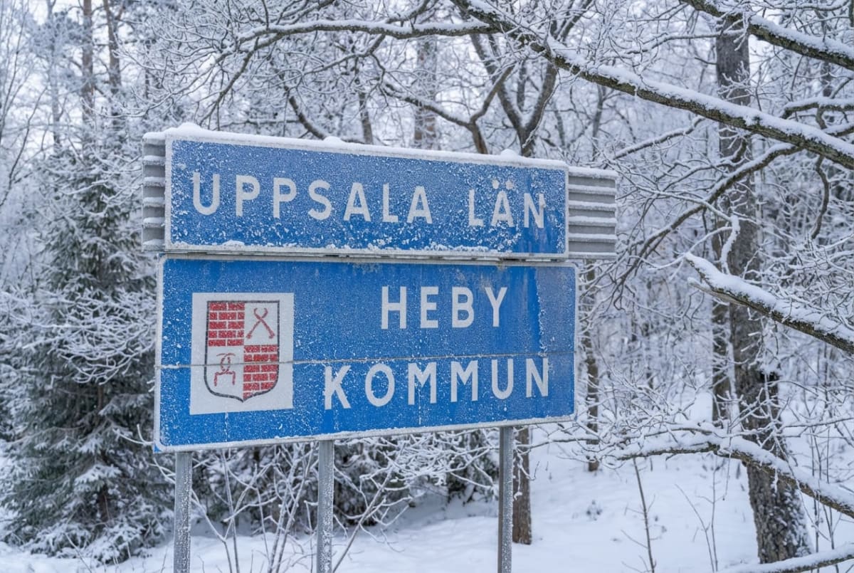 En blå skylt med texten ”UPPSALA LÄN” och ”HEBY KOMMUN” samt Heby kommuns vapen, täckt av snö och frost. Bakgrunden består av snötäckta träd och buskar i vinterlandskap.