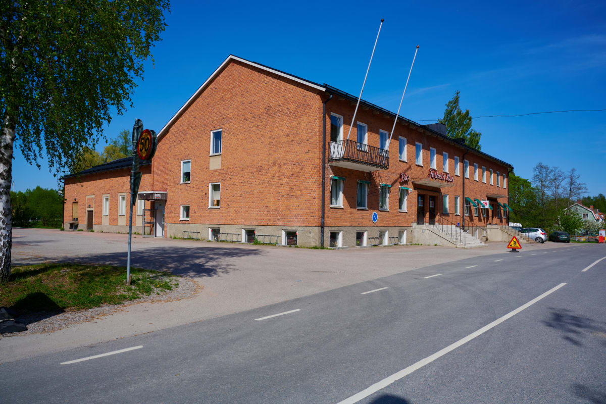 En stor röd tegelbyggnad med vita fönsterkarmar och en balkong syns under en klarblå himmel. Framför byggnaden finns en asfalterad yta och en väg med vita linjer. En hastighetsskylt med 50 km/h och en varningsskylt med ett barn på cykel syns vid vägk