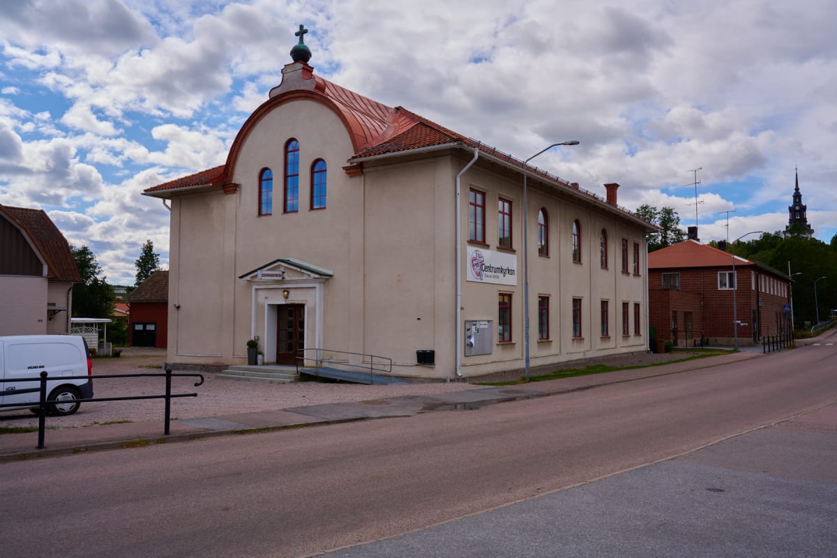 En beige byggnad med rött tak och ett kors på toppen syns på bilden. Framför byggnaden finns en väg och en trottoar. Till vänster om byggnaden står en vit skåpbil och i bakgrunden syns skymten av en kyrktorn.