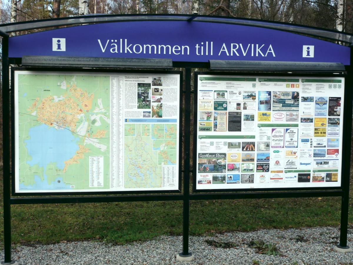En vägskylt med texten ”Välkommen till ARVIKA” framför en trädbevuxen bakgrund. Under skylten finns två informationstavlor. Den vänstra visar en karta över Arvika med omnejd. Den högra tavlan innehåller många mindre reklambilder och information om lo