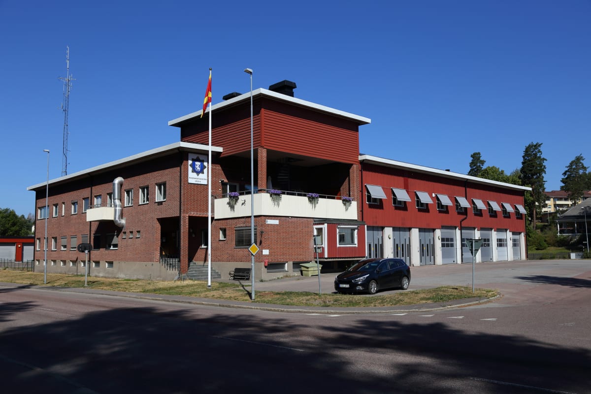 En stor, röd och tegelröd byggnad med flera garageportar och fönster står under en klarblå himmel. En svart bil är parkerad på en gräsmatta framför byggnaden. En flaggstång med en röd och gul flagga syns framför byggnaden, och i bakgrunden finns träd