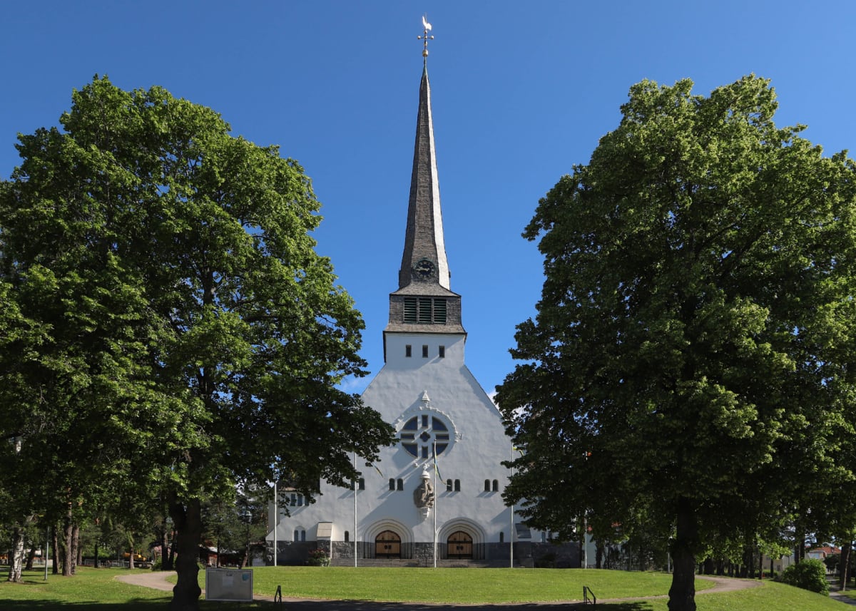 En vit kyrka med en hög spetskupolisttorn syns centralt i bilden. Två stora gröna träd ramar in kyrkan på varsin sida. Gräsmatta med en gångstig i förgrunden och blå himmel i bakgrunden.