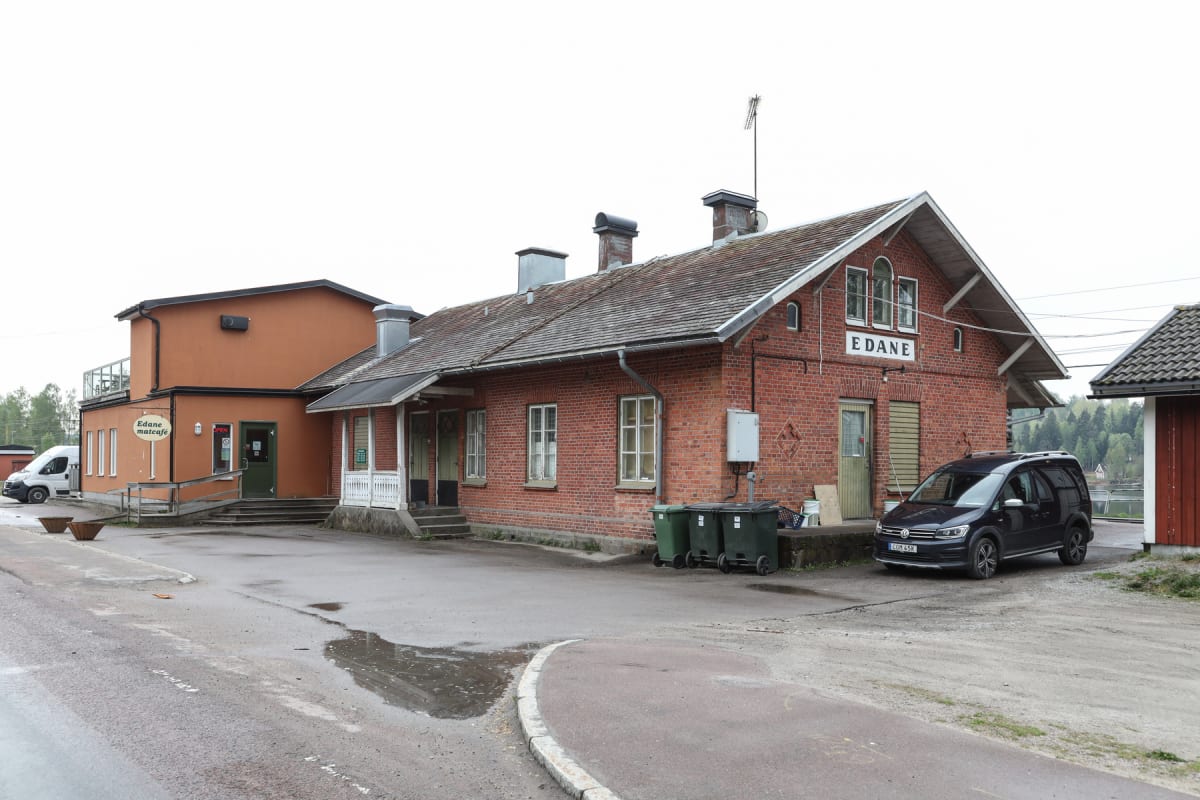 En exteriör vy av två byggnader vid en väg. Till vänster finns en orange byggnad med skylten "Edane matcafe". Till höger står en större röd tegelbyggnad med texten "EDANE" uppskyltad. Framför tegelbyggnaden står en mörk bil och tre gröna soptunnor.
