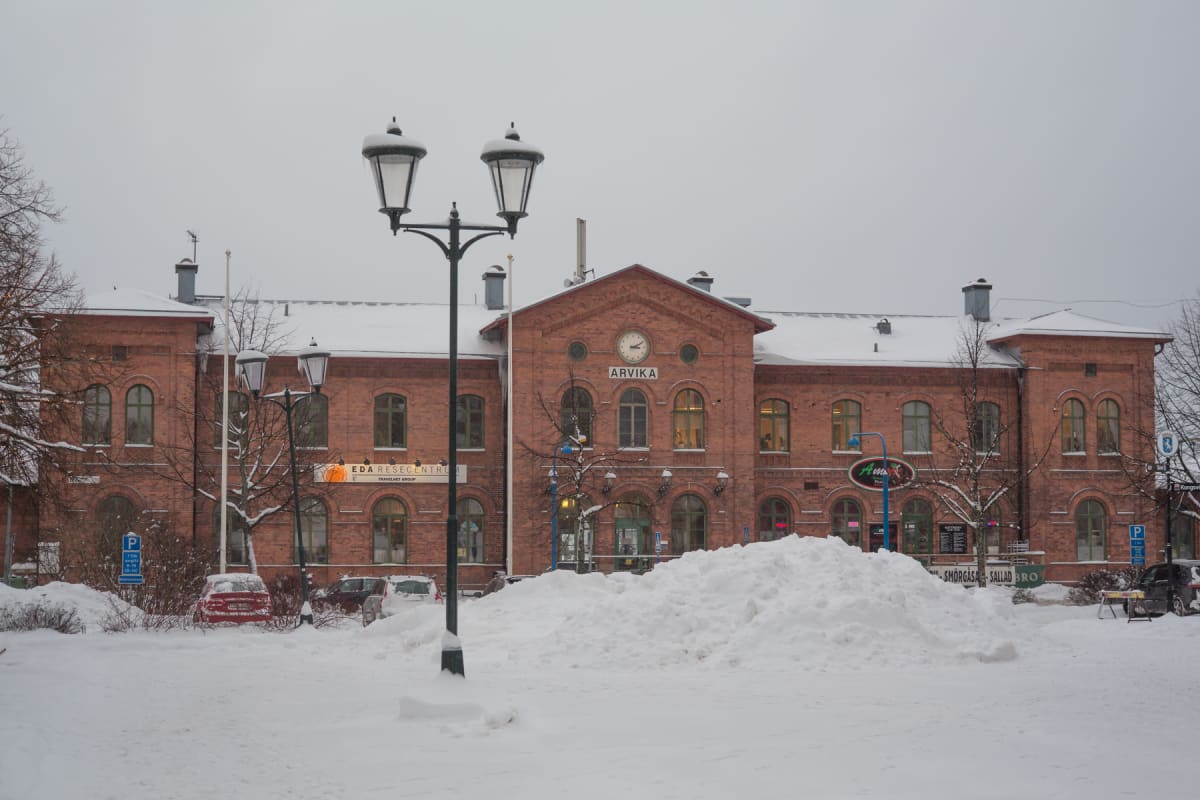 En stor, mörk lyktstolpe står i förgrunden med en byggnad i tegel bakom. Byggnaden har skylten "ARVIKA" och en klocka i mitten. Allt är täckt av snö, inklusive ett stort snöberg i förgrunden framför byggnaden. Ett par bilar syns delvis täckta av snö.