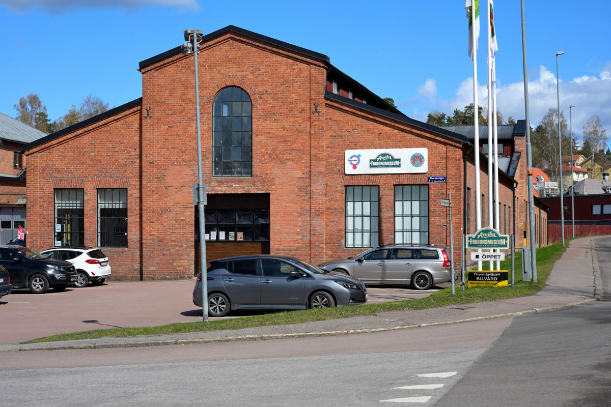 En röd tegelbyggnad med stora fönster och en logotyp för "Arvika Fordonsmuseum", parkerade bilar framför och en väg till höger. På trottoaren framför byggnaden står en skylt med texten "ÖPPET" och flera flaggstänger med flaggor.