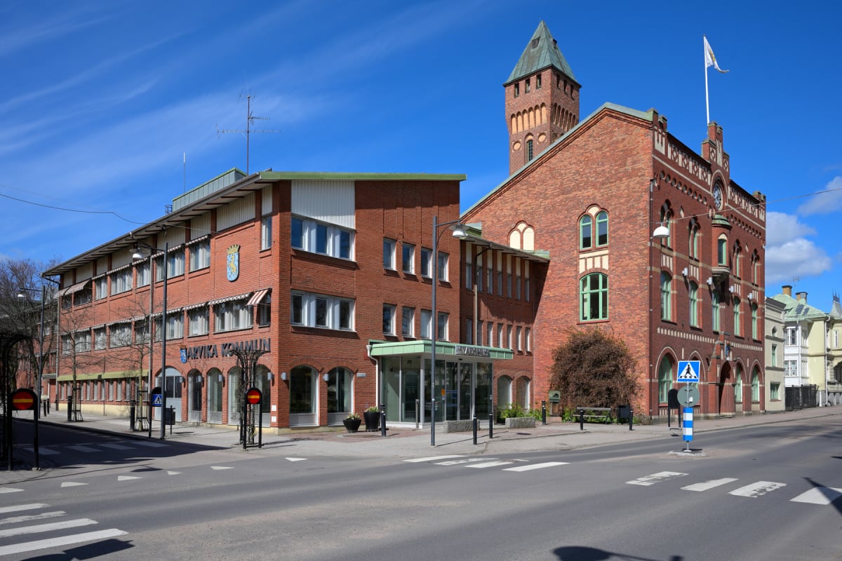 En gatuvy som visar två anslutande byggnader i tegel, varav den ena är skyltad med "ARVIKA KOMMUN". Framför byggnaderna finns en asfalterad väg med övergångsställen, och himlen är blå med några lätta moln. Till höger syns delar av fler byggnader.