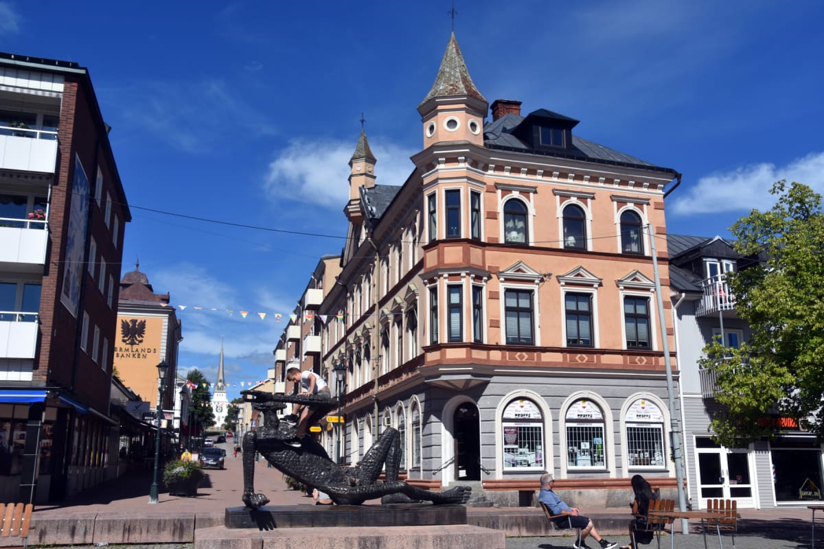 En gata i en stad med byggnader på båda sidor under en klarblå himmel. I förgrunden syns en mörk staty med en person som klättrar på den. Två personer sitter vid ett bord. Flaggor spända över gatan. En skylt med "ERMLANDS BANKEN" syns på en byggnad.