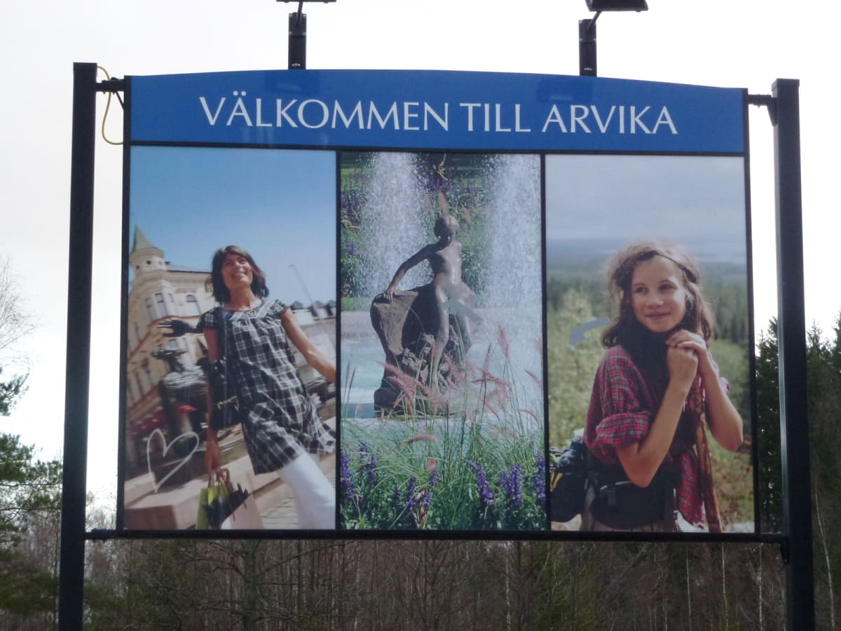 En stor blå skylt med texten "VÄLKOMMEN TILL ARVIKA" överst, under den sitter tre bilder. Den första visar en kvinna framför en byggnad, den andra visar en staty med en fontän och den tredje visar en ung kvinna med skog i bakgrunden.
