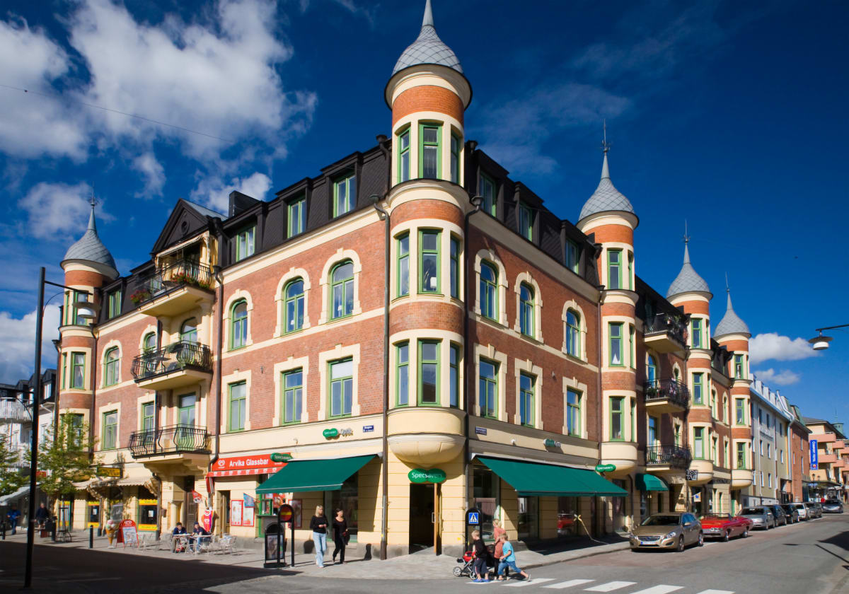 En bred bild över en stadsgata med en stor hörnbyggnad i förgrunden, parkerade bilar längs gatan, flera personer, samt en klarblå himmel med spridda moln. Byggnaden har detaljerade fönster och balkonger. En övergångsställe finns framför byggnaden