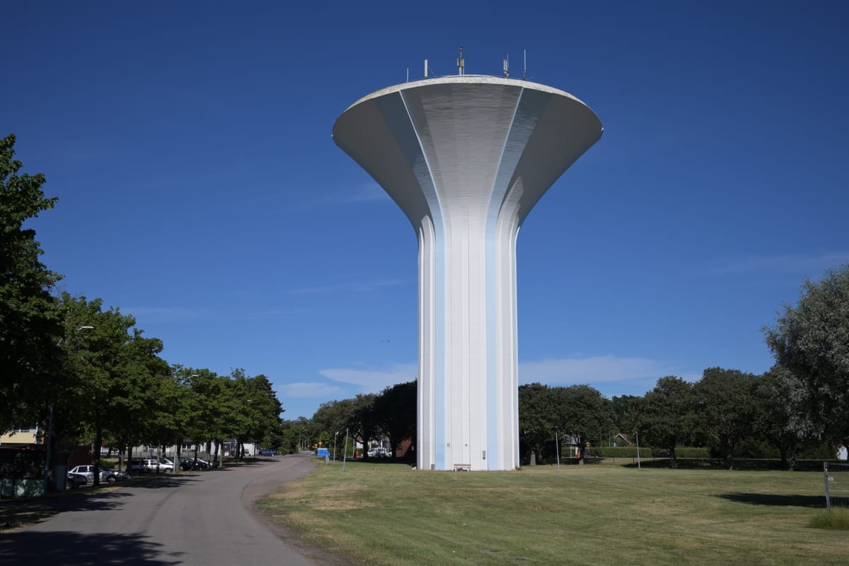 En stor, vit vattentorn med blåa vertikala ränder står i centrum under en klarblå himmel. Runt tornet finns gröna gräsmattor och lövträd. En asfalterad väg med parkerade bilar syns till vänster i bilden.