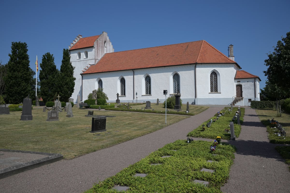 En vit kyrkobyggnad med orange tak står i centrum under en klarblå himmel. Runt kyrkan finns en gräsklädd kyrkogård med gravstenar och grusgångar, samt träd i bakgrunden.