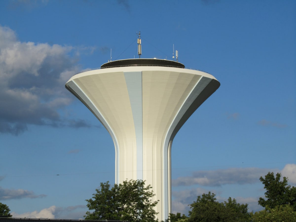 En stor, vit och beige vattenreservoar med blåa vertikala ränder under en blå himmel med vita moln. Reservoaren omges av gröna träd i förgrunden och det finns antenner på toppen av strukturen.