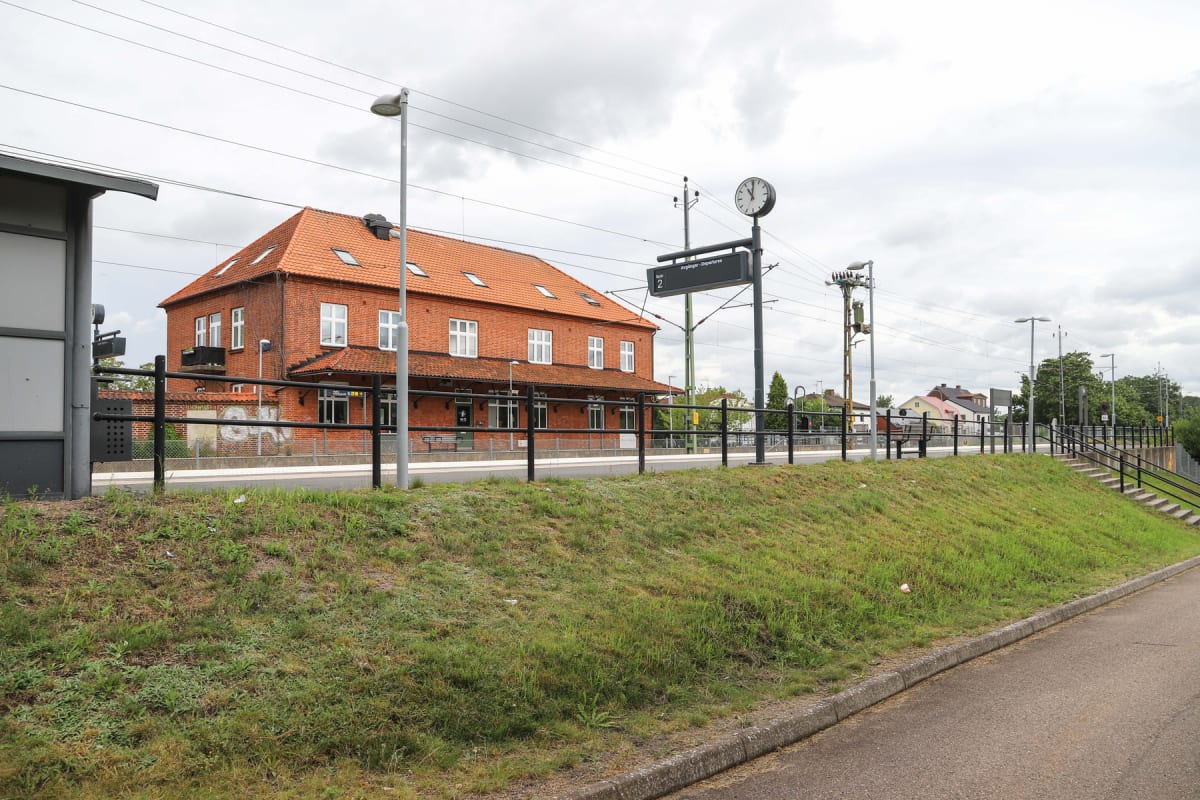 En stor röd tegelbyggnad med tegeltak syns bredvid en järnvägsstation med perrong. Framför byggnaden finns en svart metallstaket och en grön grässlänt. En klocka och en avgångstavla är synliga ovanför perrongen under en molnig himmel.
