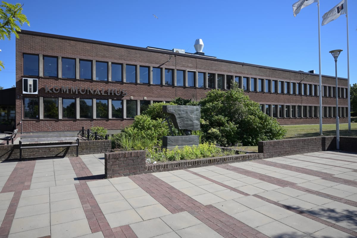 En fasad av ett kommunhus i tegel med många fönster. Framför byggnaden finns ett stenlagt torg, en trädgårdsanläggning med buskar och en skulptur, samt flaggstänger med vita flaggor som är delvis synliga. Texten "KOMMUNALHUS" syns på fasaden.