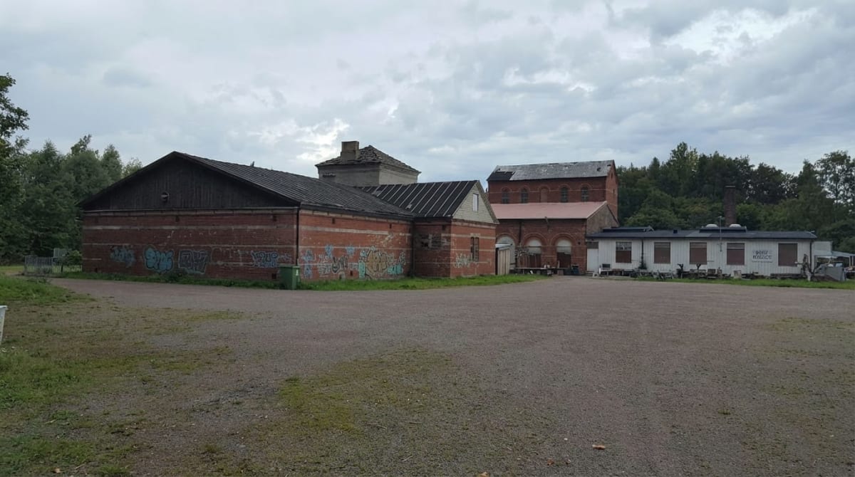 En bild som visar en grusplan framför flera byggnader med tegelväggar och svarta tak. En del av tegelväggarna är täckta med graffiti. I bakgrunden syns skog och en gråmulen himmel. En vit byggnad med platt tak och skyltningen "Borr Skånes" är också s