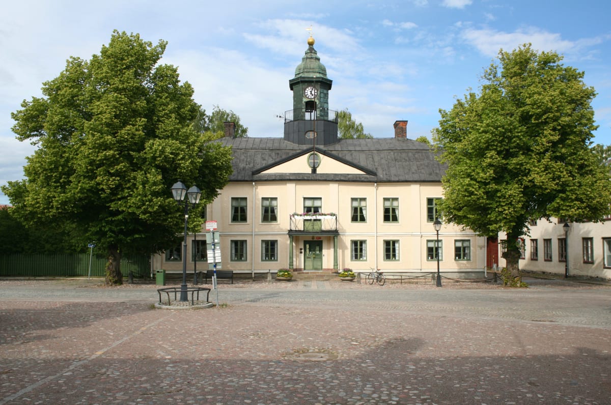 En gul byggnad med svart tak och ett koppartaksklocktorn står i centrum under en blå himmel med få moln. Framför byggnaden finns ett torg med gatsten och två stora träd på vardera sida. En cykel är parkerad vid byggnadens högra sida och gatlyktor syn