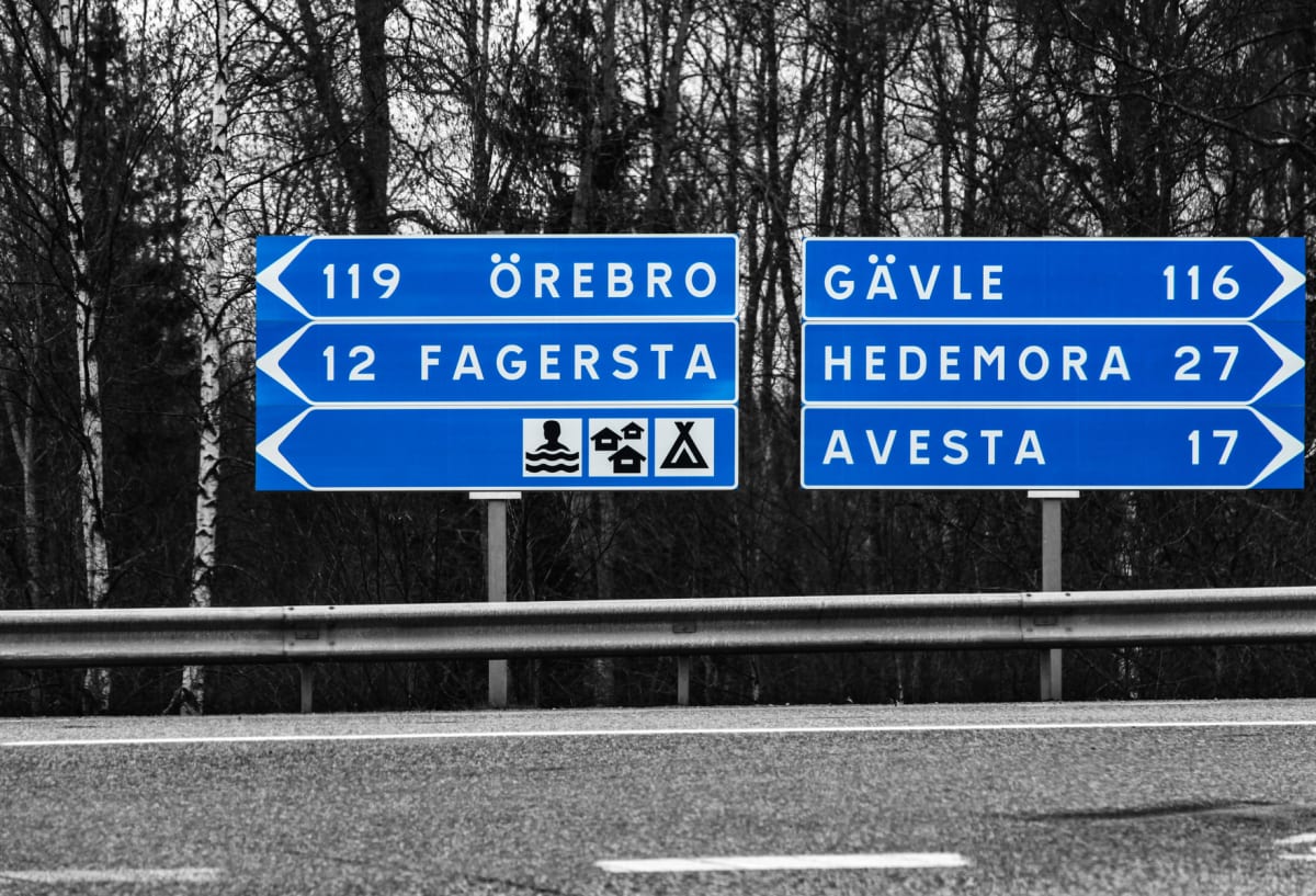En svartvit bild med färgade blå vägskyltar. Texten på skyltarna visar riktningar mot Örebro, Fagersta, Gävle, Hedemora och Avesta, samt symboler för badplats, camping och tältplats. I bakgrunden syns träd utan löv.