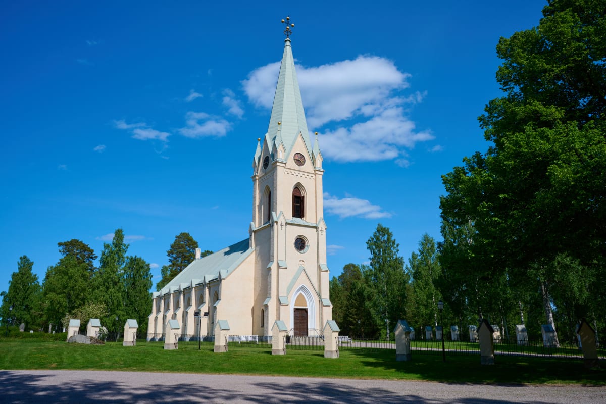 En gul kyrka med ett högt, grått spetsigt tak och torn under en klarblå himmel med vita moln. Kyrkan är omgiven av grön gräsmatta, lummiga träd och vad som verkar vara en kyrkogård med stenar med små tak. En grusväg syns i förgrunden.