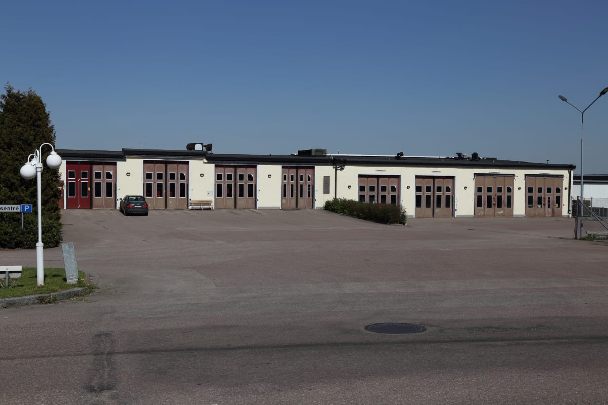 En fasad med flera portar i rött och brunt, parkeringsplats framför, en mörk personbil parkerad, och en lampstolpe med en P-skylt syns till vänster. Byggnaden har en ljusgul färg.