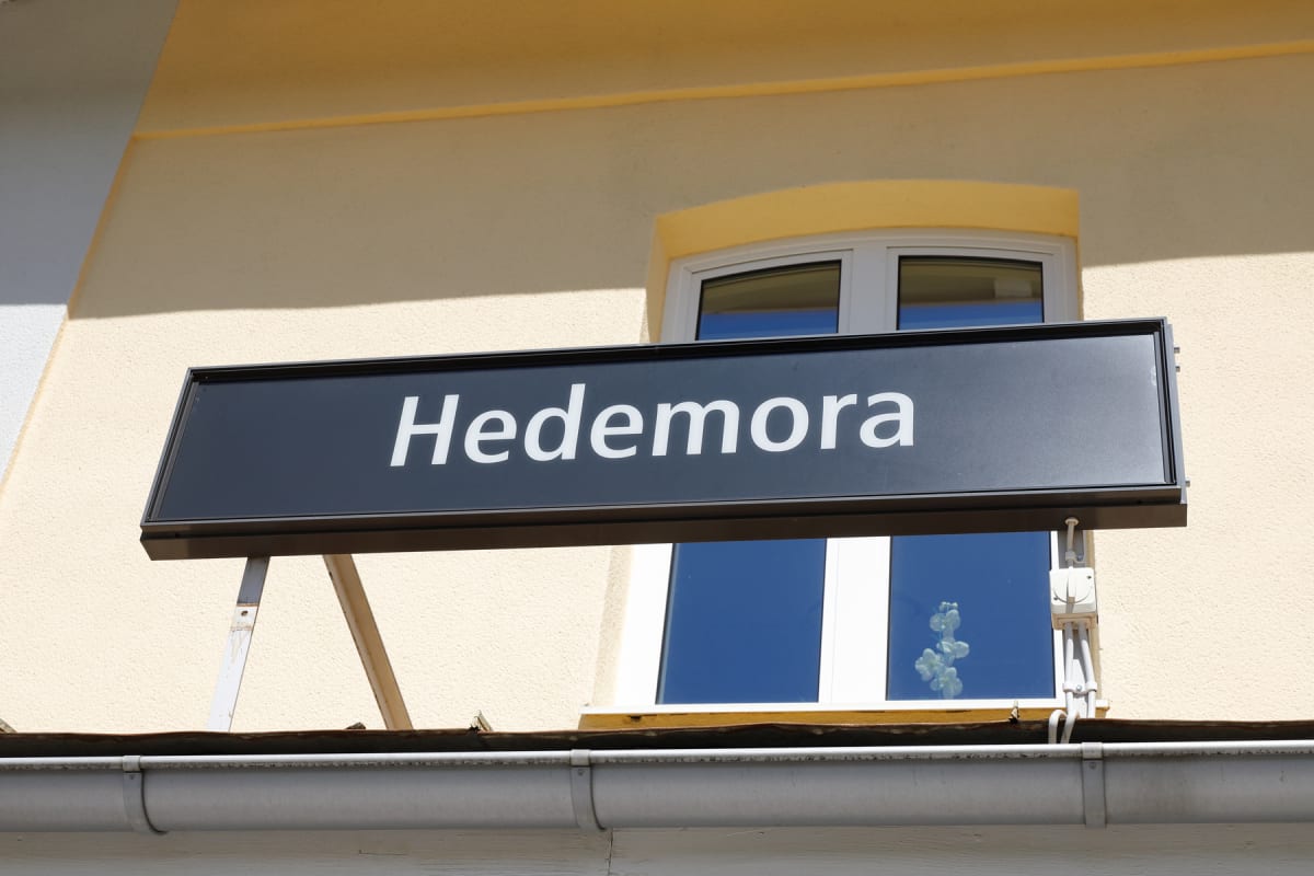 En svart skylt med vit text "Hedemora" är monterad på en ljusgul byggnadsfasad. Under skylten syns ett fönsterparti med blå himmel speglandes i rutorna, samt en takränna nedtill i bilden.