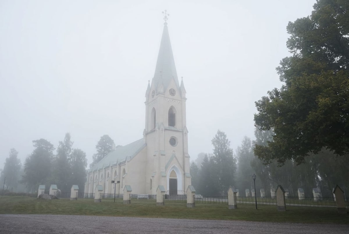 En gulvit kyrkobyggnad med spetsigt grönt tak och torn syns i dimma. Byggnaden är omringad av grönska, inklusive träd och gräsmattor, samt en kyrkogård med flertalet gravstenar. En grusväg löper i förgrunden.