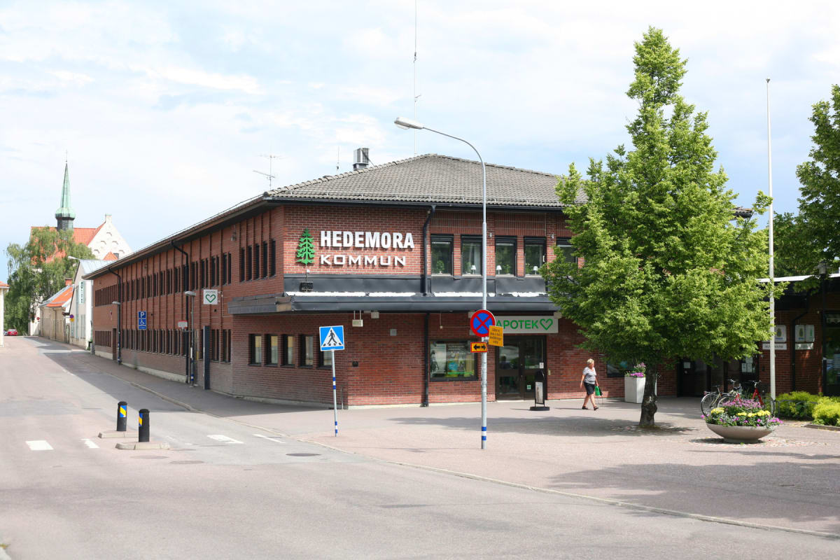En bild tagen utomhus från en gata som leder in mot två byggnader. Till vänster syns flera tegelbyggnader, varav den största har texten "HEDEMORA KOMMUN" och en grön trädlogotyp på fasaden. Till höger om den byggnaden skymtar "APOTEK"-skylten. En per