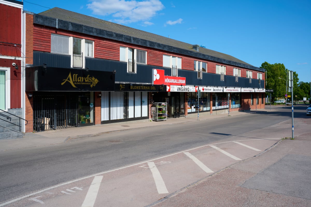 En gathörna med en lång byggnad med röd fasad och svarta detaljer, där flera butiker är belägna på bottenvåningen. På gatan finns en cykelbana och en bil som kör där. Blå himmel med moln ses ovanför byggnaden.