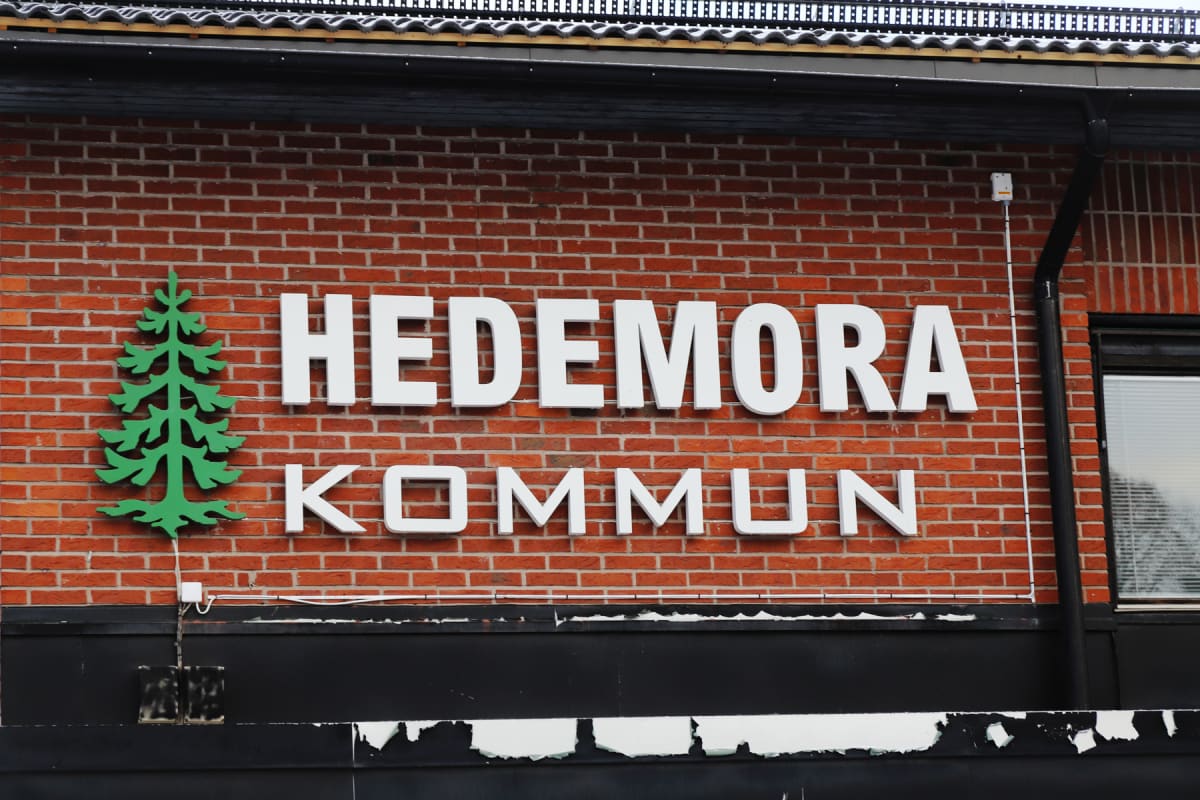 En fasad av rött tegel med vita bokstäver som bildar texten "HEDEMORA KOMMUN". Till vänster om texten finns en grön logotyp föreställande en stiliserad gran. Ovanför texten syns en takkant med stupränna och svarta takpannor, och nedanför finns en sva