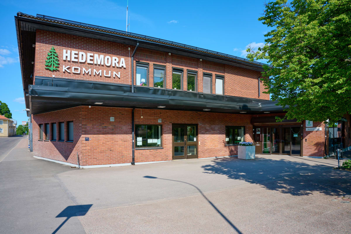 En fasadvy av Hedemora kommunhus med en stor grön logotyp av en gran och texten "HEDEMORA KOMMUN" på den röda tegelväggen. Ingången till byggnaden är synlig med dubbeldörrar och det står en blomlåda framför. Till höger skymtar ett träd.