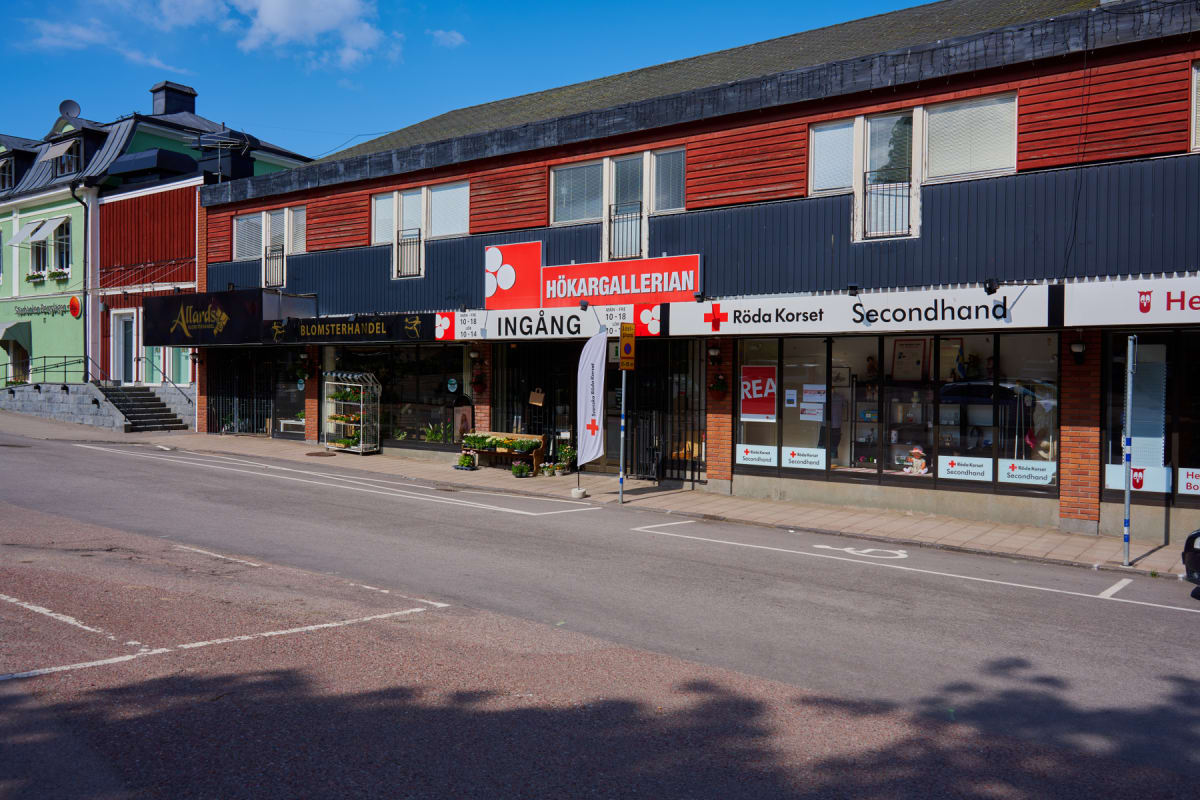 En bild visar en gata framför en rad byggnader med butiker på bottenvåningen. Byggnaderna har röda och gröna fasader med mörkblå detaljer. Flera butiksskyltar syns, inklusive "HÖKARGALLERIAN", "BLOMSTERHANDEL" och "Röda Korset Secondhand".