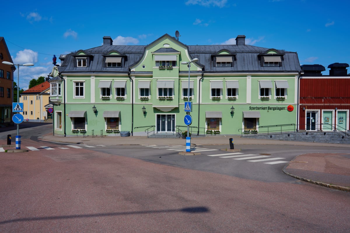 En gathörna med en ljusgrön flervåningsbyggnad med många fönster och en mörkgrå taklist, samt en del av en röd byggnad till höger. På trottoaren framför byggnaden står trafikskyltar, en gatlyktstolpe och det finns ett övergångsställe på gatan.