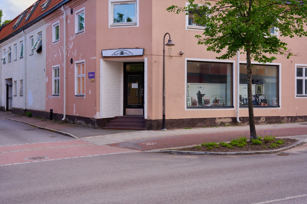 En gathörna med en två etage rosa byggnad som har butiksfönster på bottenvåningen. Bredvid den rosa byggnaden syns en vit byggnad med röda takpannor. En gatlampa står vid hörnet och en trädplantering med ett träd syns framför byggnadens fönster. Gatm