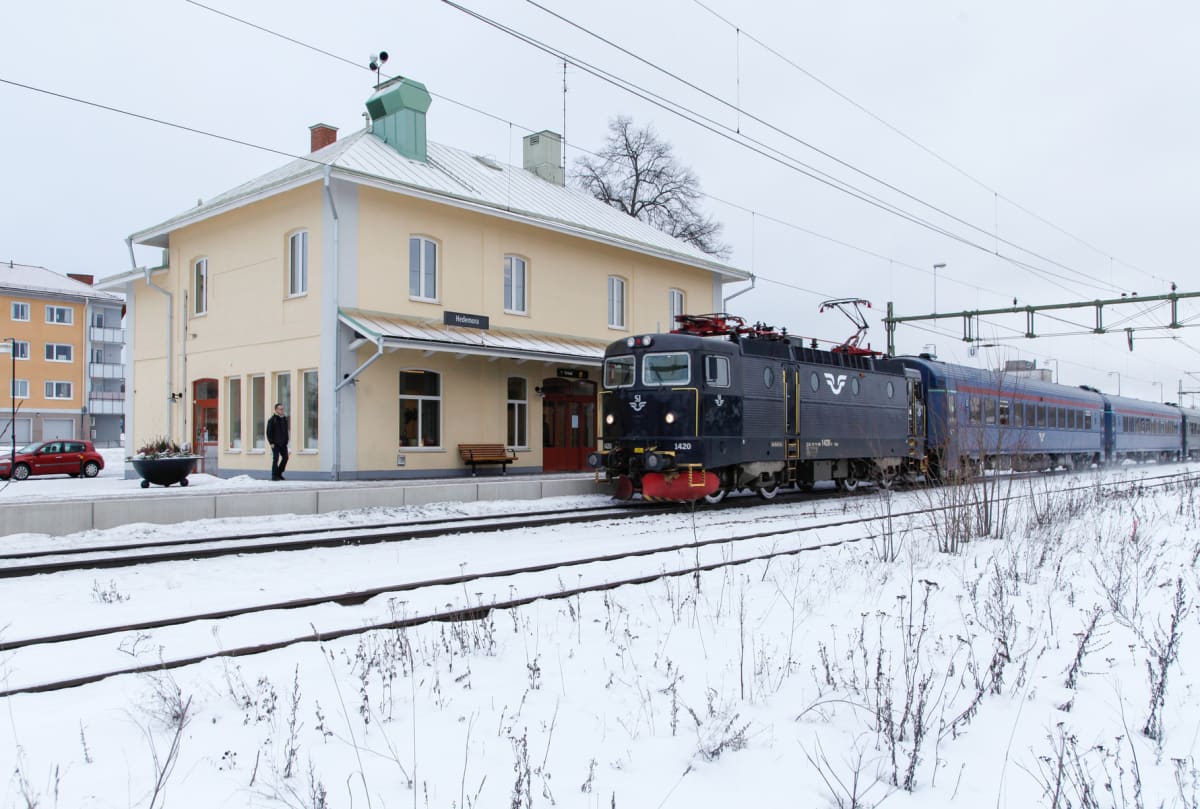 En svart SJ-lokomotiv med vagnar står vid ett ljust gult stationshus i snöigt vinterlandskap. Skylten på stationshuset visar ortsnamnet 'Hedemora'. En person står på perrongen, och en röd bil är parkerad i bakgrunden.