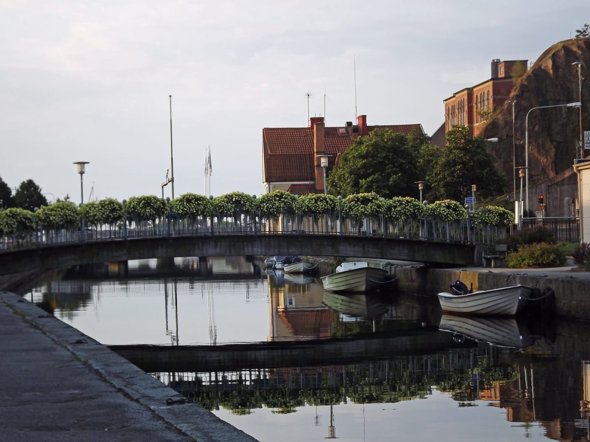 En bro med metallräcken och blomlådor sträcker sig över en kanal med stilla vatten. Flera småbåtar ligger förtöjda längs kanalbanken. I bakgrunden syns hus med röda tegeltak och en bergvägg. Reflektioner av bron och byggnader syns i vattnet under en
