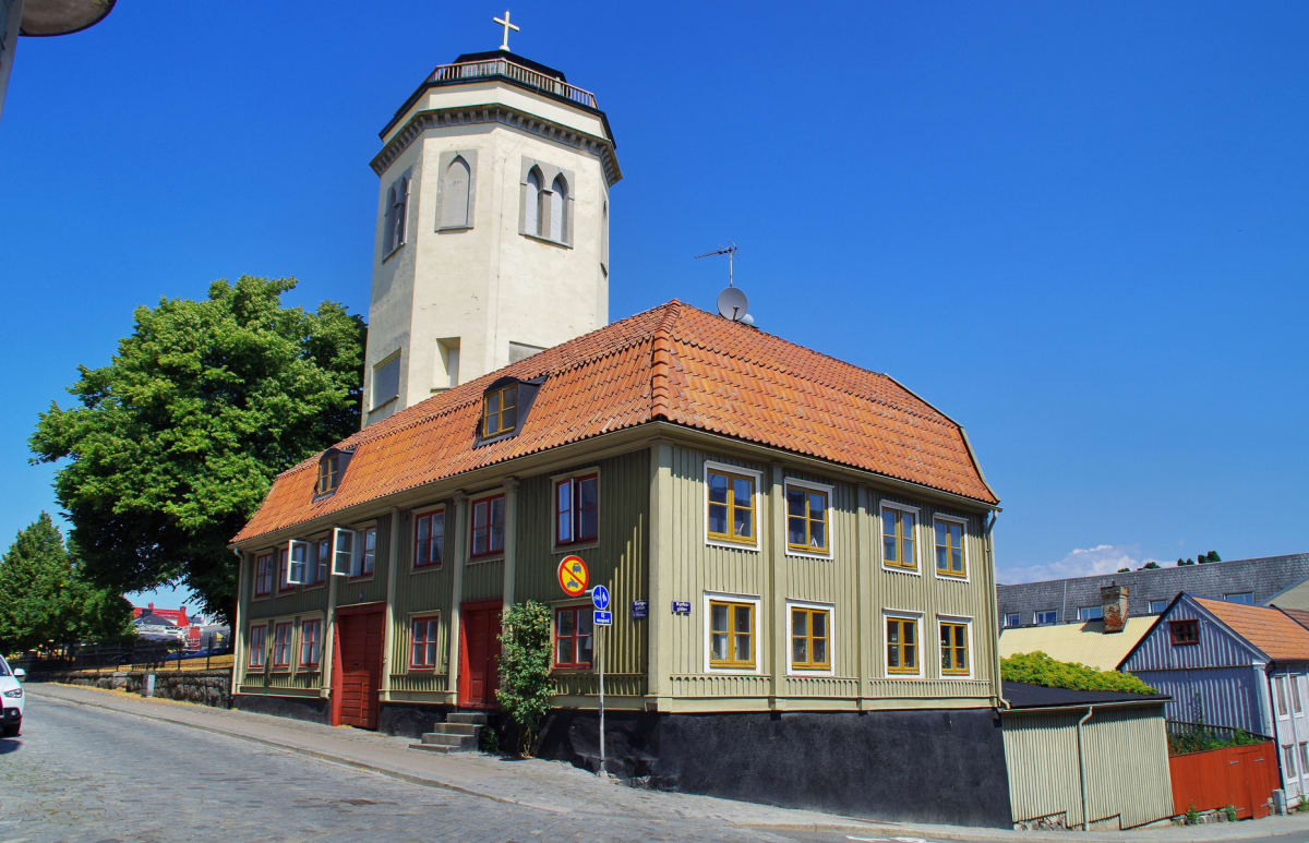 En bild som visar en ljusgrön träbyggnad med rött tegeltak och gula fönster, med en vit tornbyggnad som syns bakom. Blå himmel, gröna träd och en kullerstensgata framför byggnaden, samt andra hus och en del av en bil i bakgrunden.