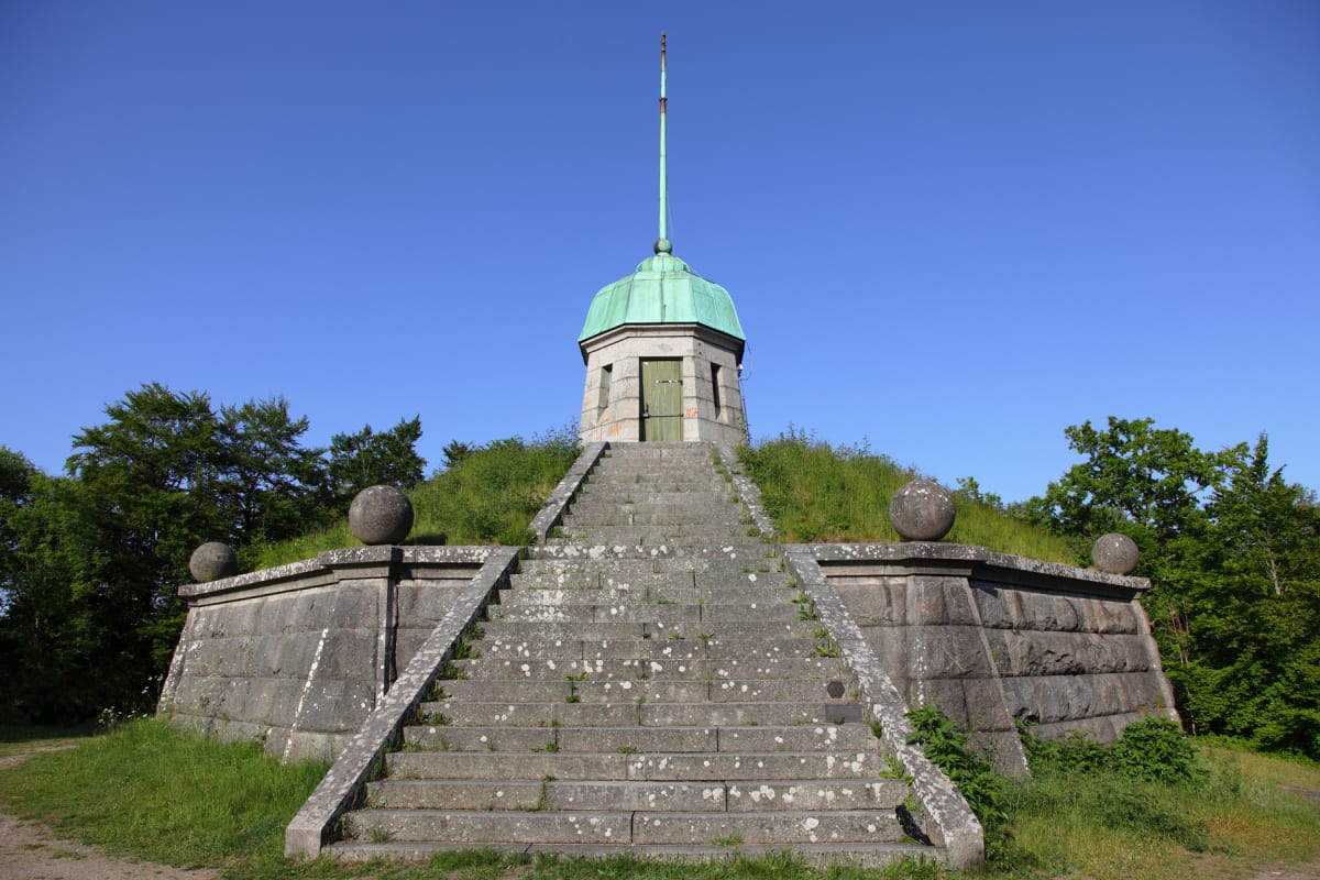 En stenbyggnad med grön kupol och en spira på toppen, uppåtsträvande trappa och stenmur. Byggnaden är omgiven av gräs och lummiga träd, allt under en klarblå himmel. Två stenklot syns på varsin sida av trappan.