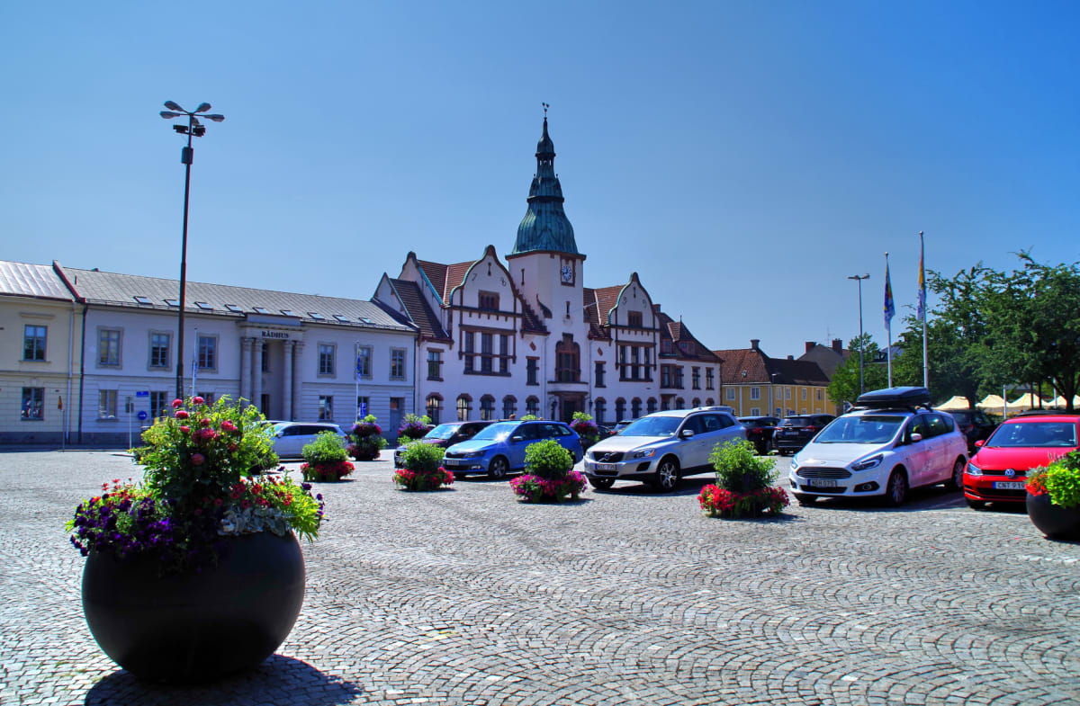 En stor översiktsbild av ett torg med en kullerstensbelagd yta. I bakgrunden syns flera byggnader, inklusive en med ett högt, grönt torn och en skylt där det står "RÅDHUS". Ett antal bilar är parkerade på torget, och flera stora blomkrukor med färggl