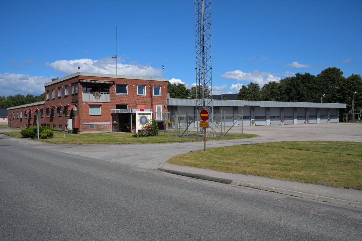 En bild som visar en brandstation med ett tvåvånings bostadshus i tegel framför flera garageportar. En metallmast står till höger om bostadshuset. Framför fastigheten finns en asfalterad yta med gräsytor vid sidorna.