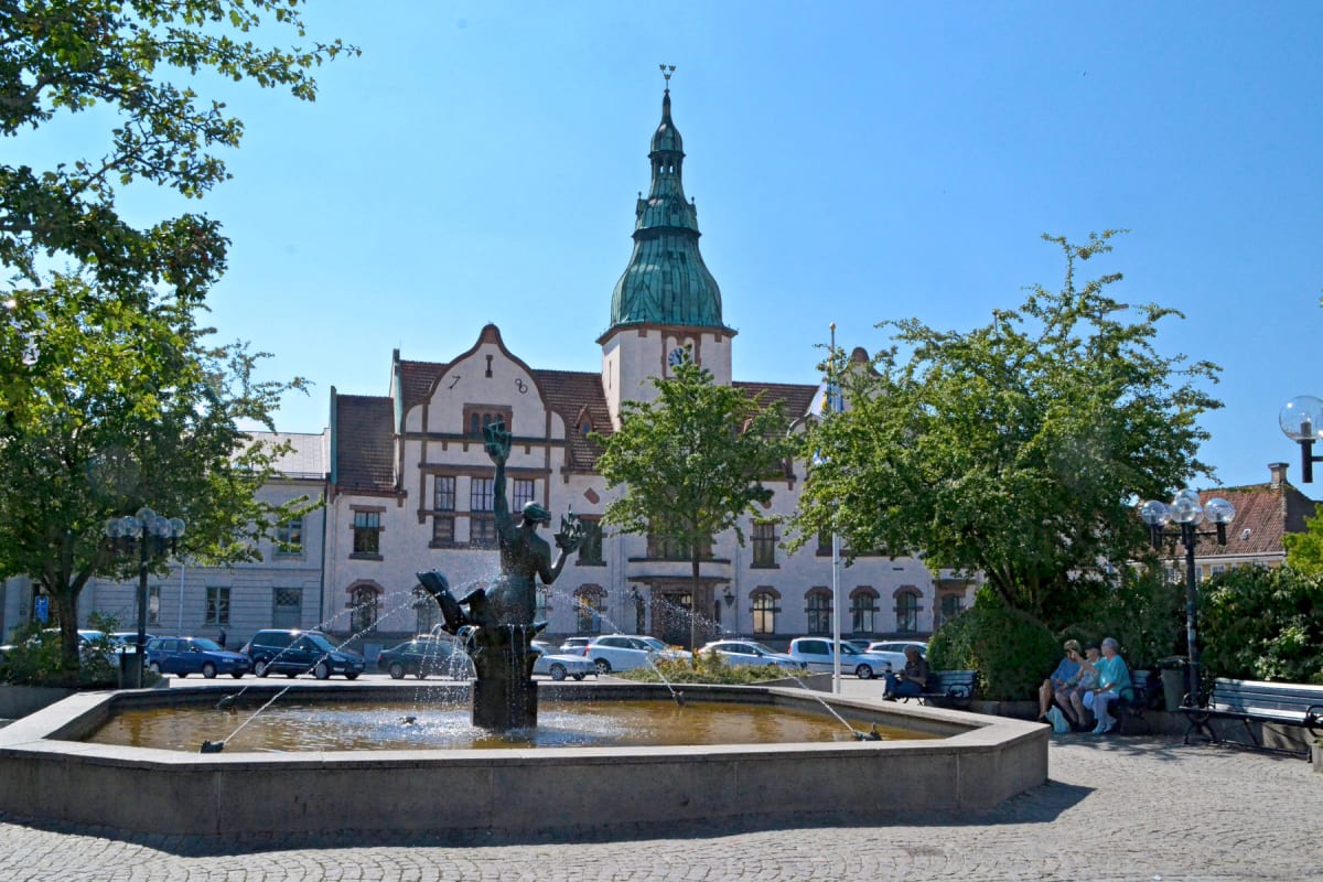 En bild som visar en fontän med en skulptur i förgrunden. I bakgrunden syns en större byggnad med ett torn och en klocka. Flera människor sitter på parkbänkar till höger om fontänen, och bilar är parkerade längs gatan bakom fontänen. Bilden är tagen