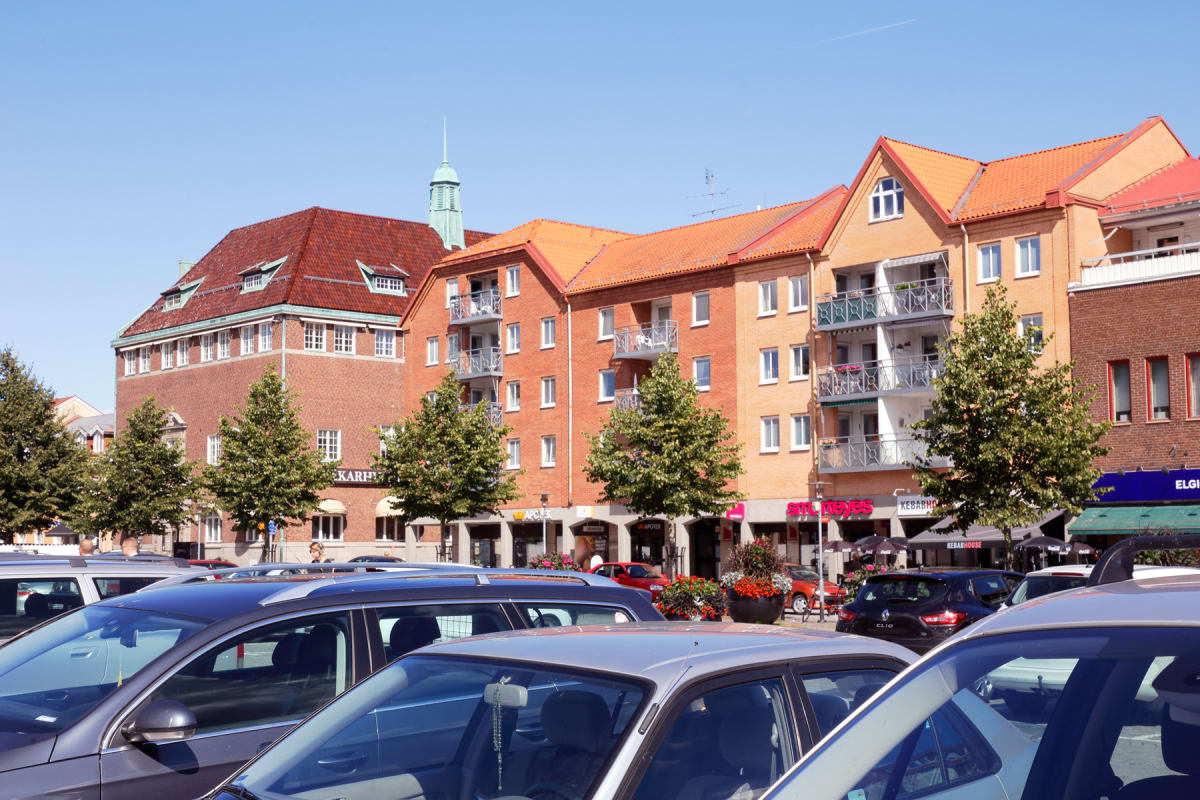 En översiktsbild av en stadsmiljö med parkerade bilar i förgrunden och byggnader med tegelfasader och röda tegeltak i bakgrunden. Mellan bilarna och byggnaderna syns träd planterade längs en gata.