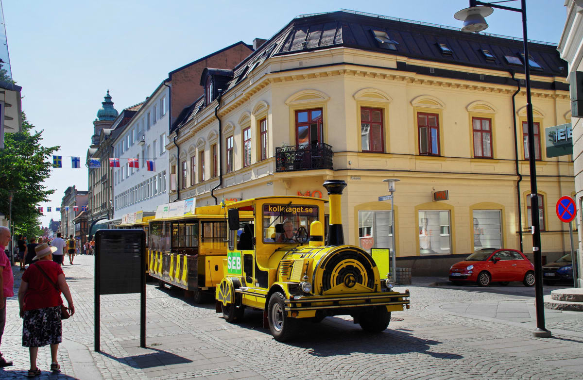 En gul vagn dragen av en traktorliknande enhet, med texten "kollekaraget.se" och "SEB", kör på en kullerstensgata. Byggnader i ljusa färger, en del med balkonger, kantar gatan. Flaggor hänger tvärs över gatan, och det finns människor på trottoaren.