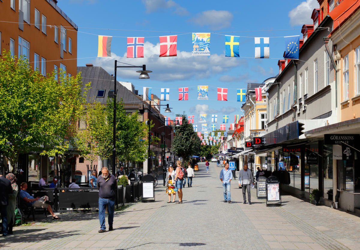 En gata med byggnader på båda sidor under soligt väder. Över gatan hänger rader med flaggor från flera nordiska länder, EU och en skylt för Karlshamn. Människor promenerar eller sitter vid uteserveringar. Träd med gröna löv ramar in bilden.