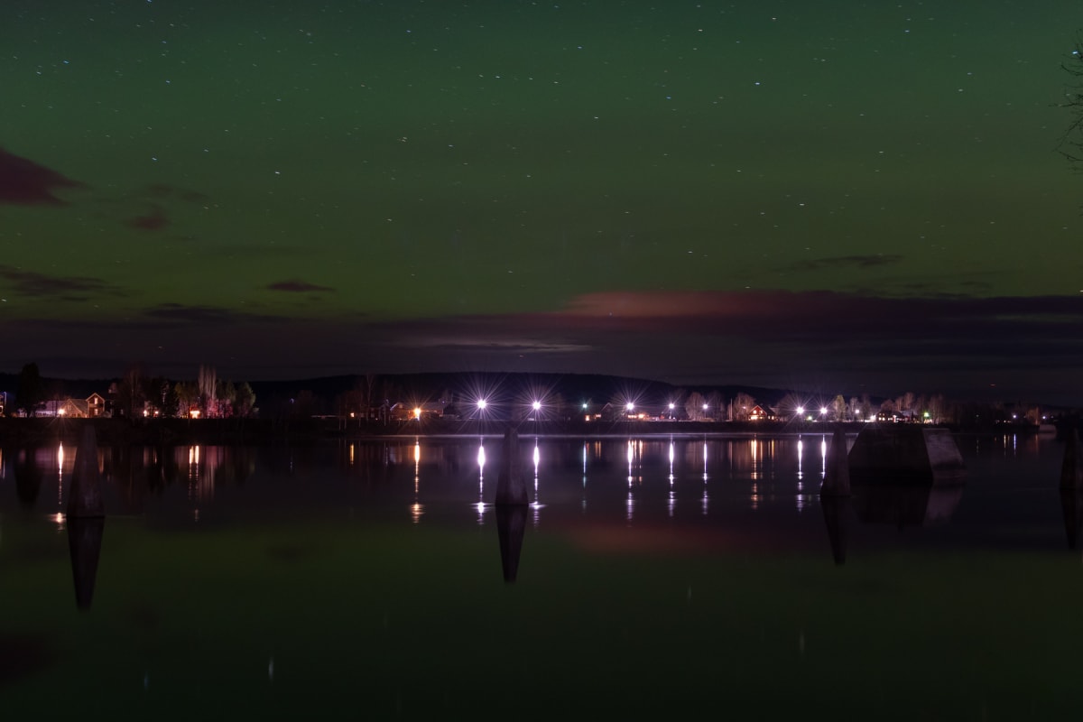 En nattbild som visar en grönfärgad himmel med stjärnor och svaga moln över ett vattenområde. På bortre stranden syns en rad upplysta byggnader och träd, reflekterade i vattnet. I förgrunden syns också mörka strukturer i vattnet.