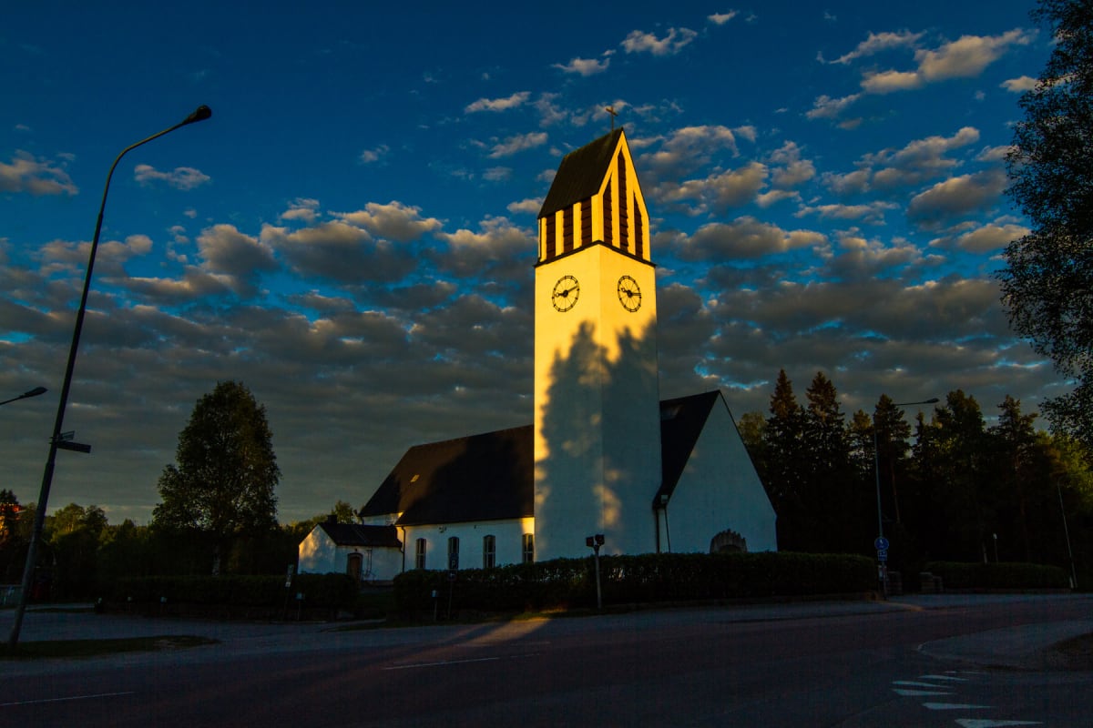 En vit kyrka med en framträdande klocktorn i gult och svart står under en blå himmel med moln. En gatlykta syns i förgrunden till vänster, och träd omger kyrkan. Solen kastar långa skuggor över tornet och marken, vilket indikerar att det är kvällssol