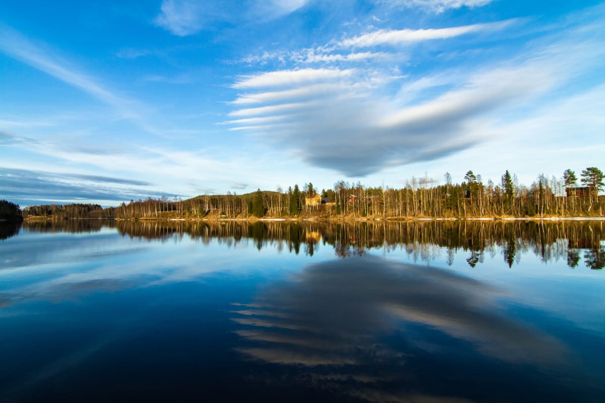 En bild som visar en stilla sjö med reflektioner av den omgivande naturen och molnen. I bakgrunden syns en skogsklädd strandlinje med barr- och lövträd samt några byggnader. Himlen är blå med vita moln som reflekteras i vattnet.