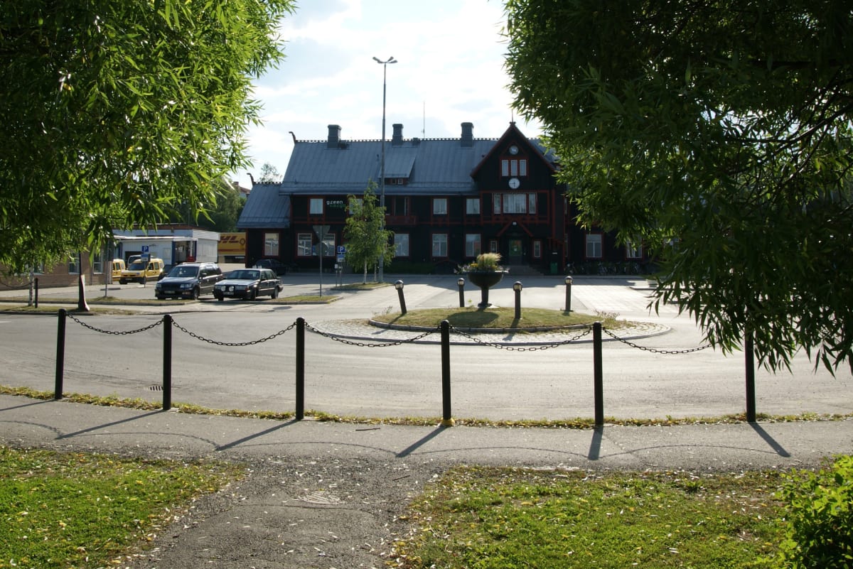 En bild tagen utomhus genom lövverk som ramar in en gata med en central byggnad i bakgrunden. Tre bilar är parkerade till vänster om byggnaden, och en liten rondell med en blomma finns framför den. Hela scenen ramas in av lövverk från träd på sidorna