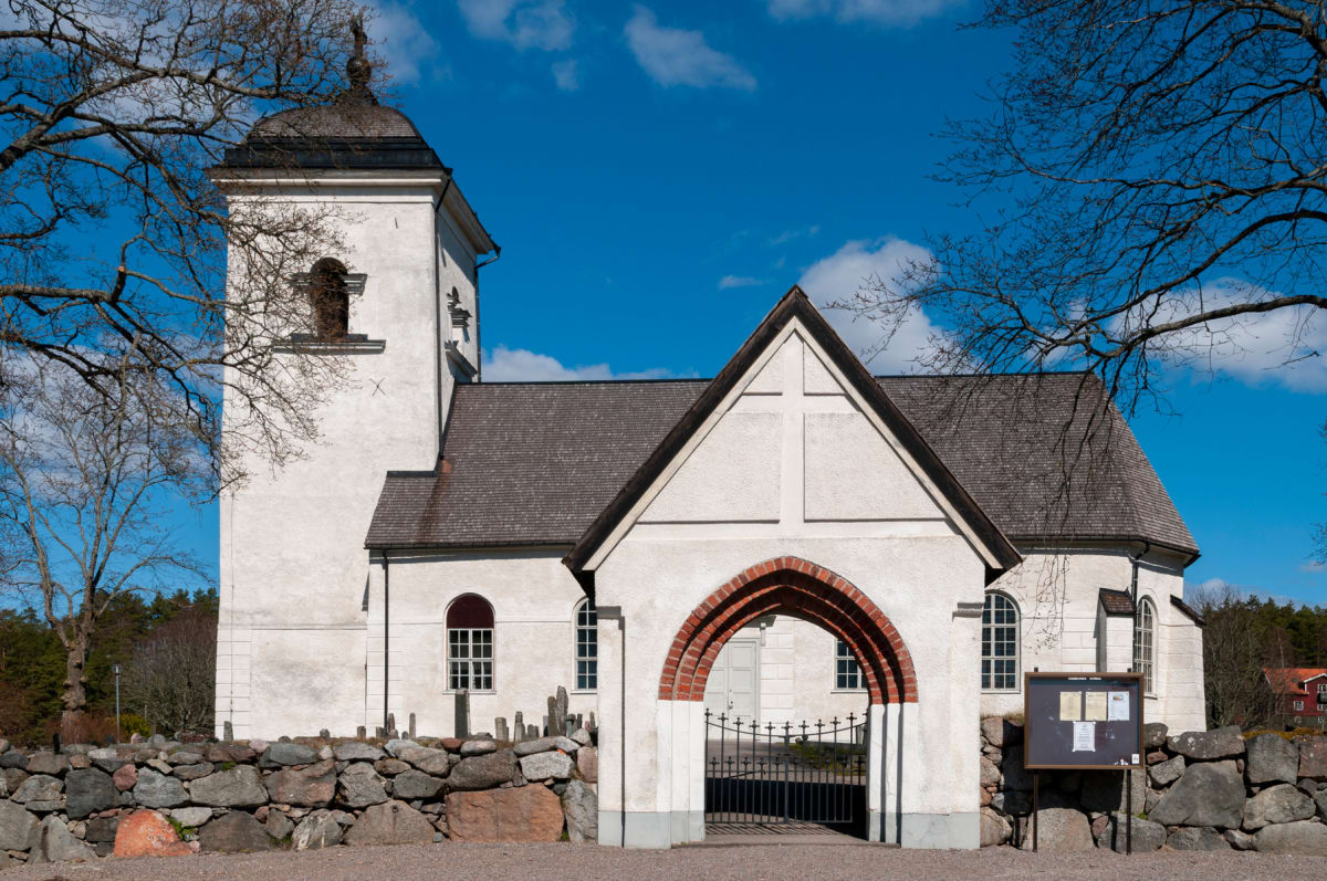 En vit kyrka med ett brunt tak och en kyrktorn i förgrunden, omgiven av kala trägrenar under en blå himmel med några moln. En stenmur syns i förgrunden, och en svart grind under en portal i tegel leder in till kyrkogården.