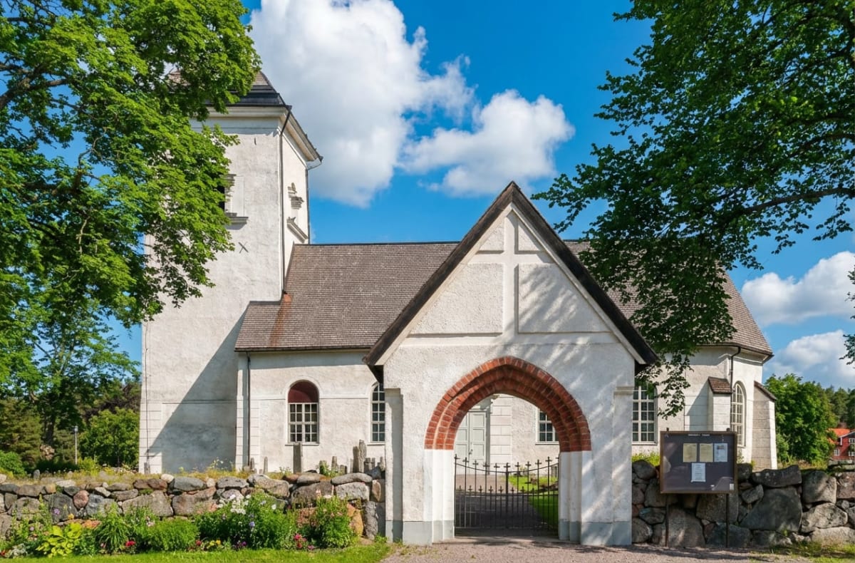 En vit kyrkobyggnad med grått tak under en blå himmel med vita moln. Framför kyrkan finns en stenmur med planteringar och en portik med ett valv av rött tegel. Till vänster och höger om kyrkan syns gröna träd.