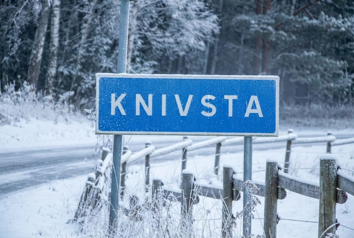 En blå skylt med texten "KNIVSTA" i stora vita bokstäver är placerad vid sidan av en snöig väg. Runtomkring skylten finns snöklädda träd, buskar och ett staket i trä. Marken och träden är täckta av snö och frost, vilket tyder på vinterväder.